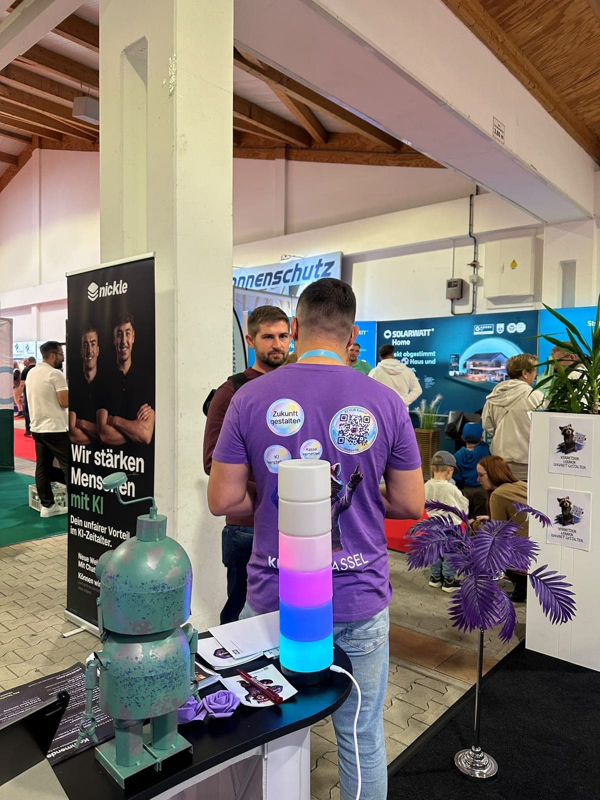 Rückenansicht eines Mannes im lila KI-Hub-Kassel-Shirt mit Aufklebern und QR-Code im Gespräch mit einem Besucher, vorne eine leuchtende Säule und eine türkise Roboterfigur.