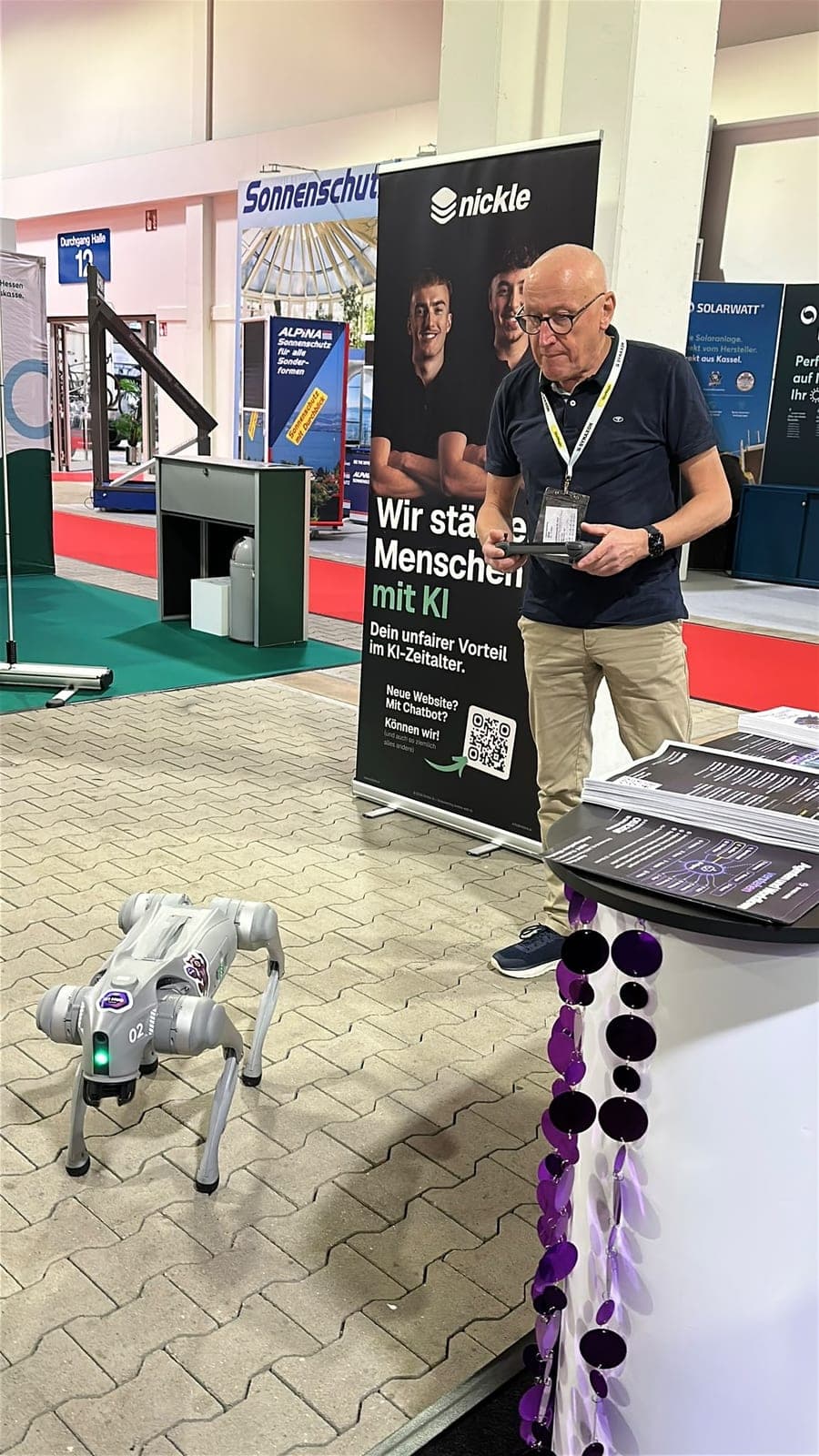 Ein Mann mit Controller steuert einen grauen Roboterhund auf dem gepflasterten Hallenboden neben dem Stand.