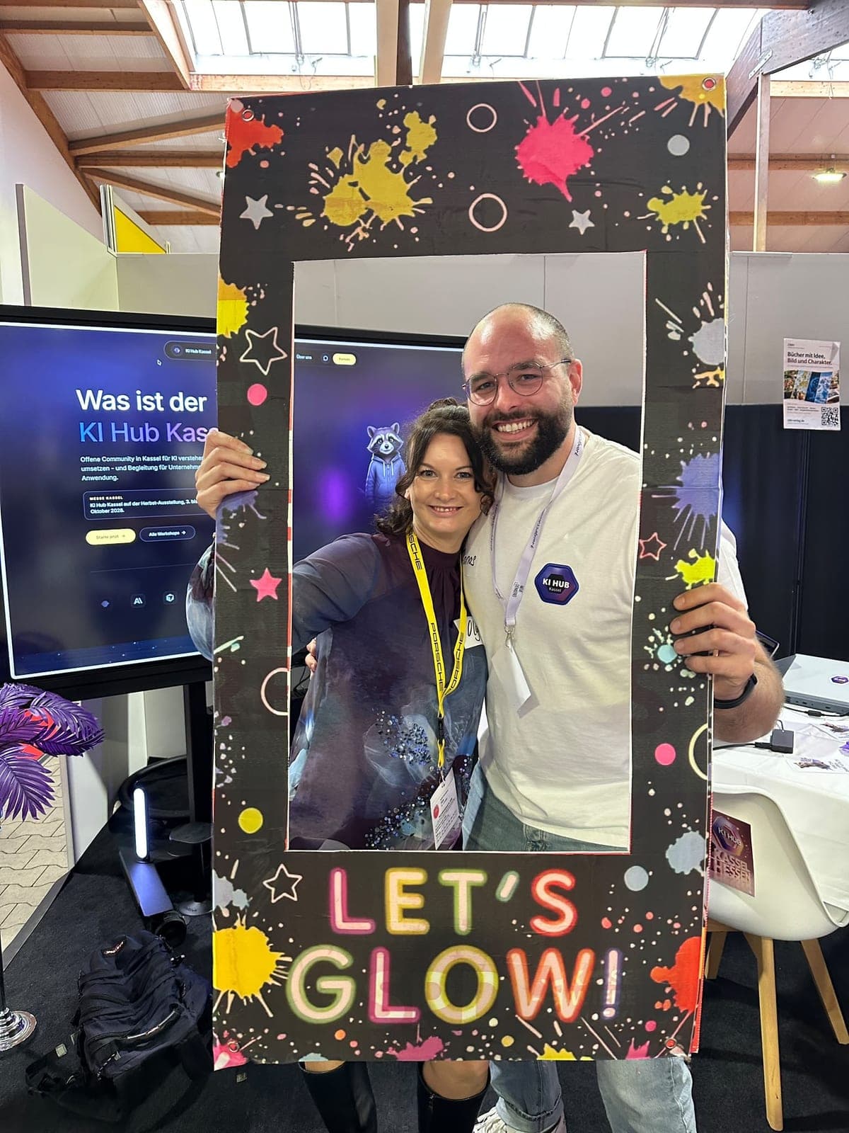 Eine Frau und ein Mann halten lächelnd einen bunten Fotorahmen mit der Aufschrift „Let’s Glow!“, dahinter ein Bildschirm mit der KI-Hub-Kassel-Website und dem Waschbär-Maskottchen.
