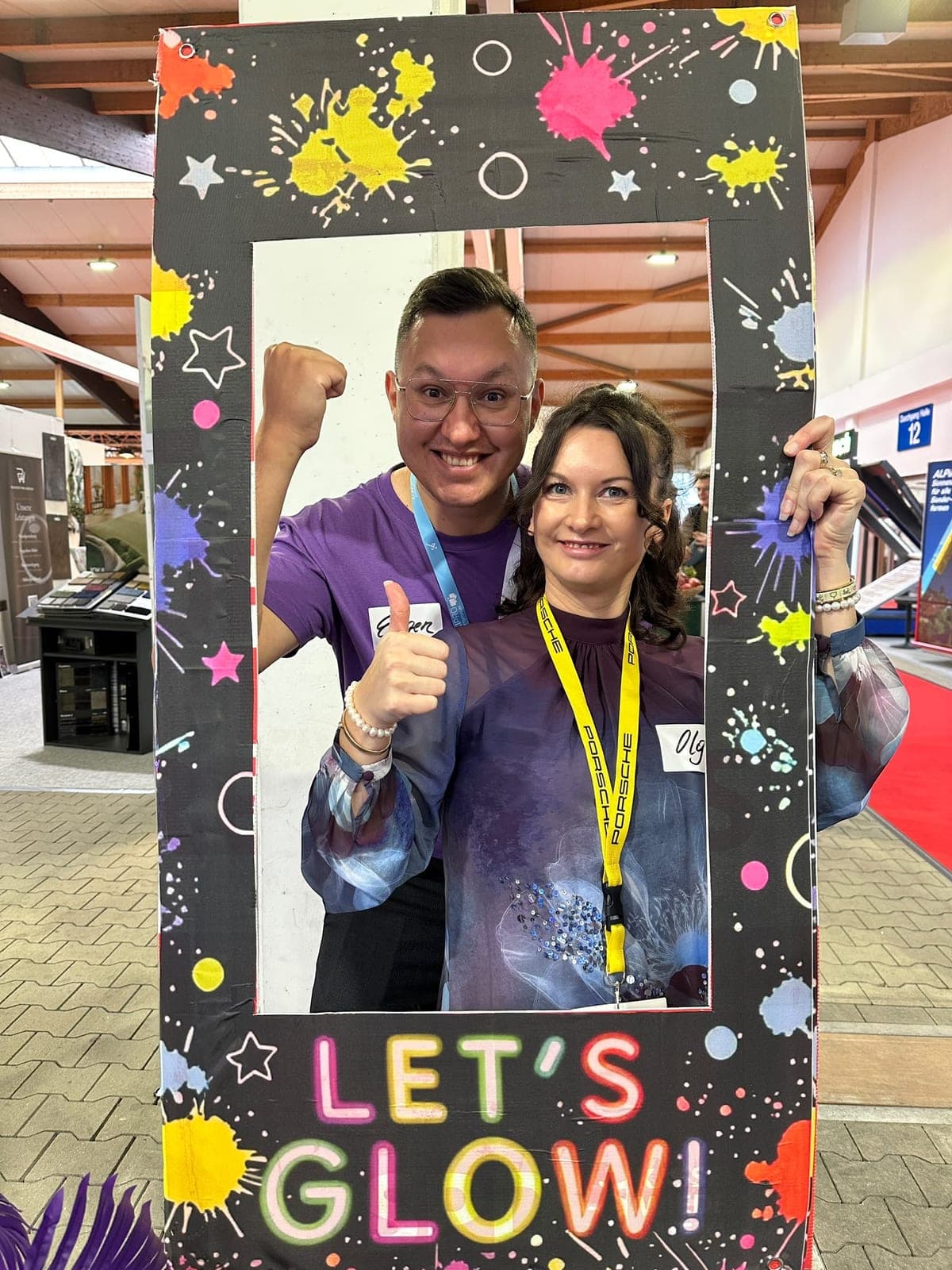 Ein Mann im lila KI-Hub-Shirt und eine Frau posieren mit erhobener Faust und Daumen hoch im „Let’s Glow!“-Fotorahmen in der Messehalle.