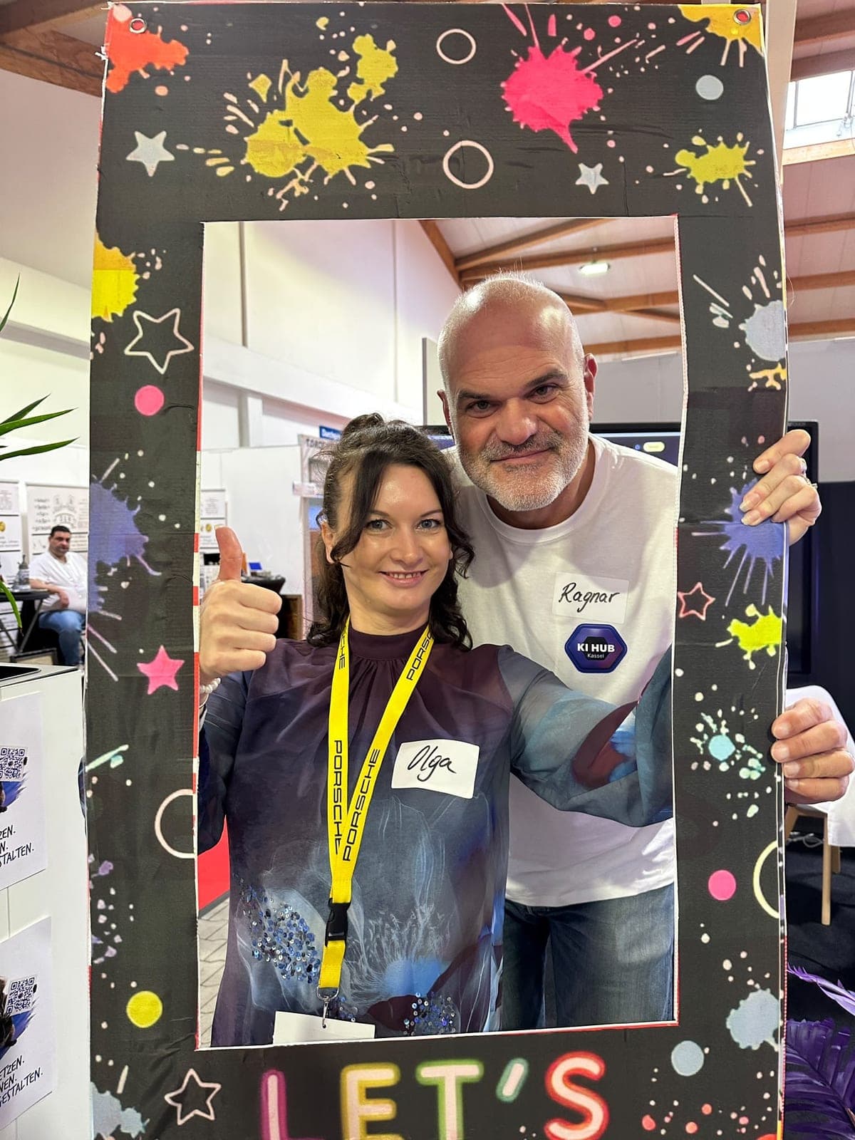 Eine Frau mit Daumen hoch und ein Mann im weißen KI-Hub-Shirt posieren im „Let’s Glow!“-Fotorahmen am Stand.